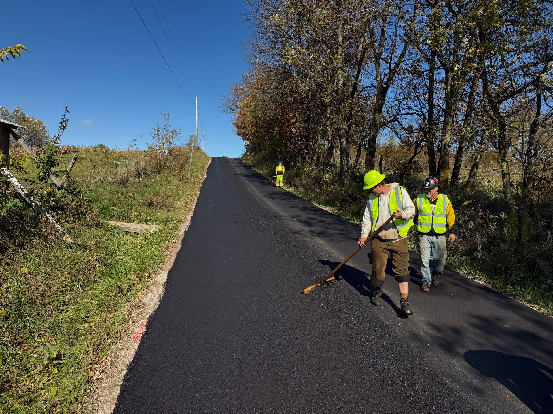 Roadworkers in high-visibility vests repaving a rural road on a sunny day