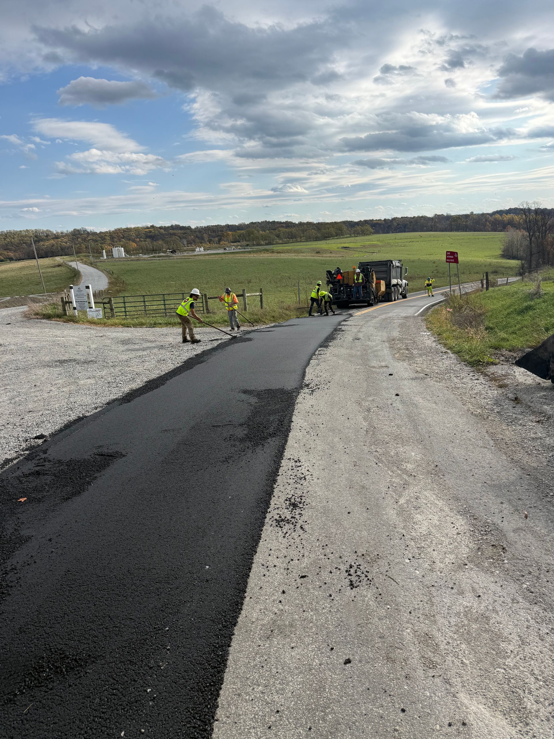 Road paving crew laying fresh black asphalt on a gravel rural road under a cloudy sky