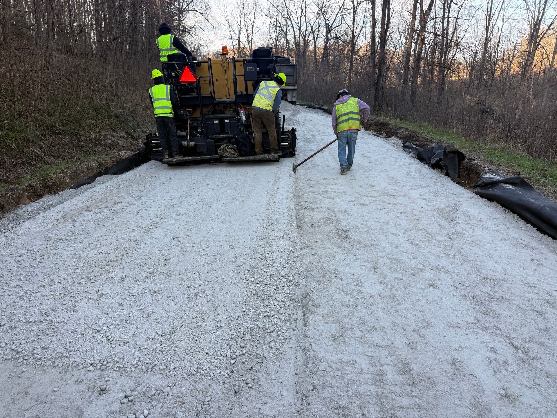Workers paving a rural road with a paver and fresh gravel, one worker walking ahead.