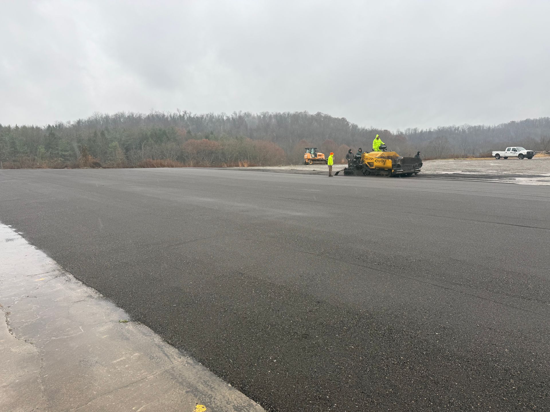 Wet runway with workers and a yellow construction vehicle near a wooded shoreline under cloudy skies