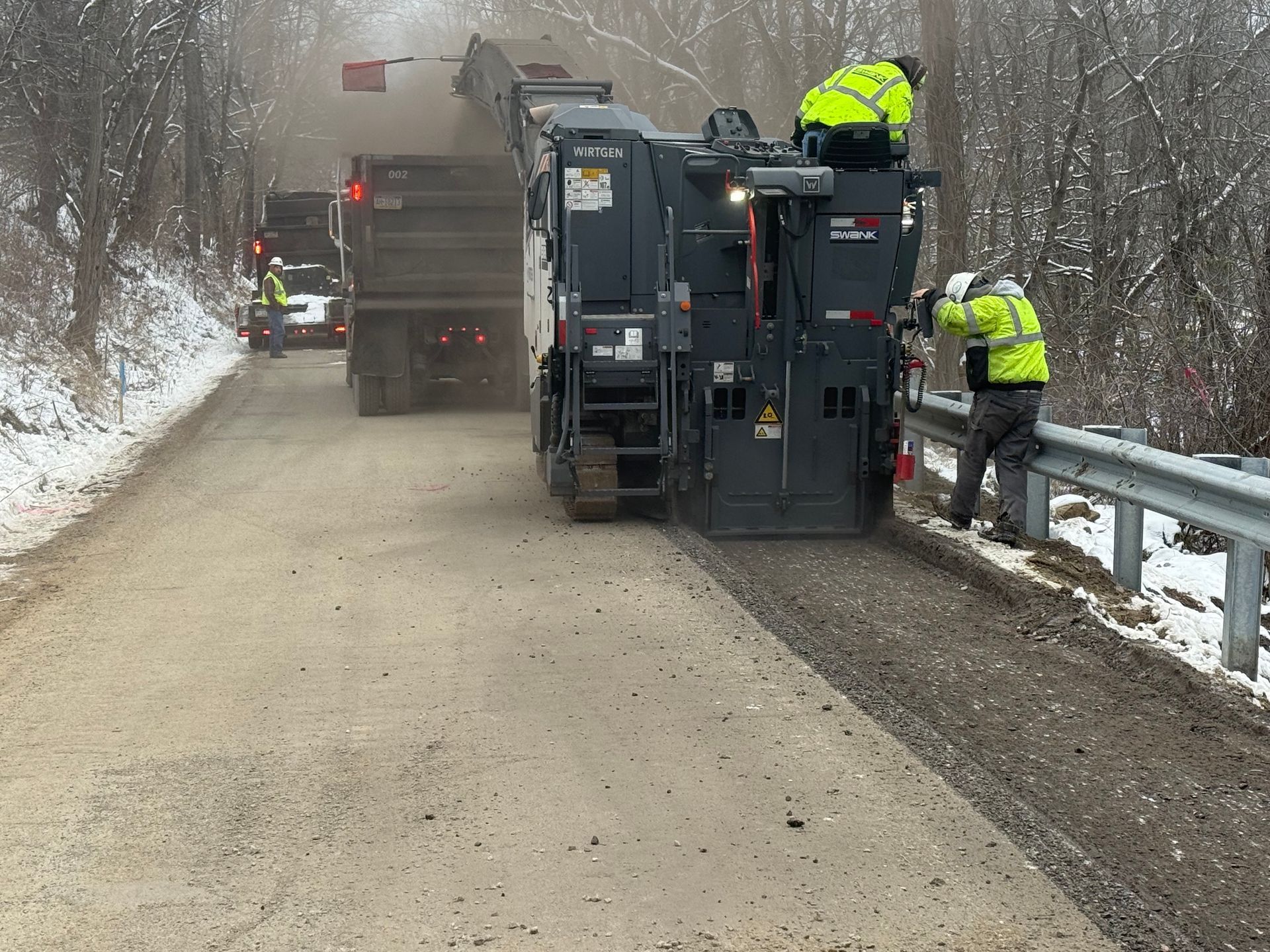 Road crew paving a snowy roadside, with heavy machinery and workers in neon jackets.