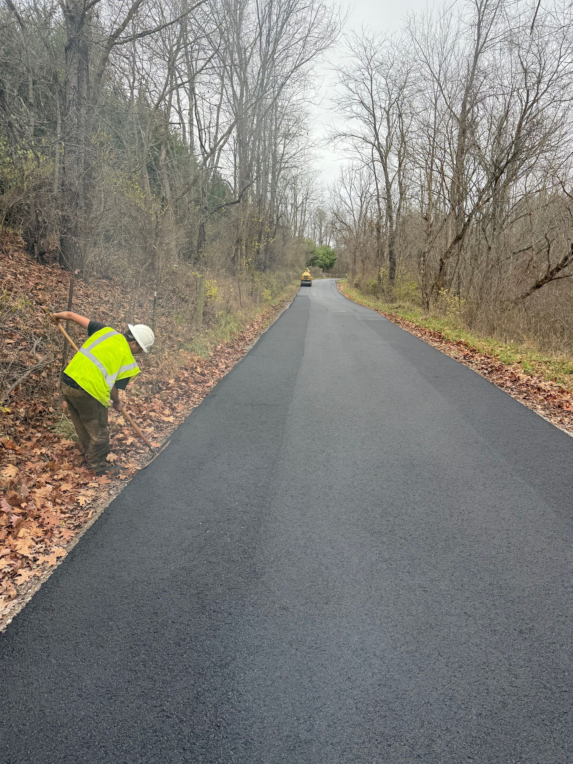 Person in a neon jacket working beside a newly paved rural road lined with leafless trees.
