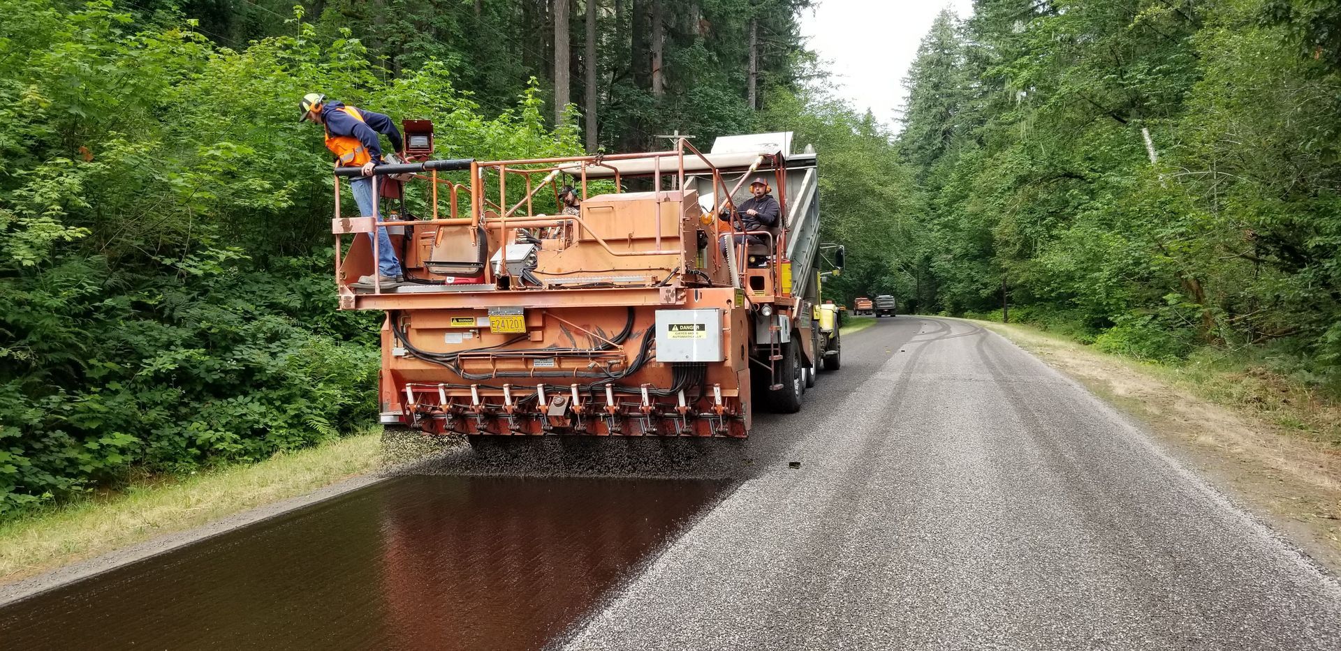 Orange road maintenance truck spreading fresh asphalt on a gravel road beside dense green trees