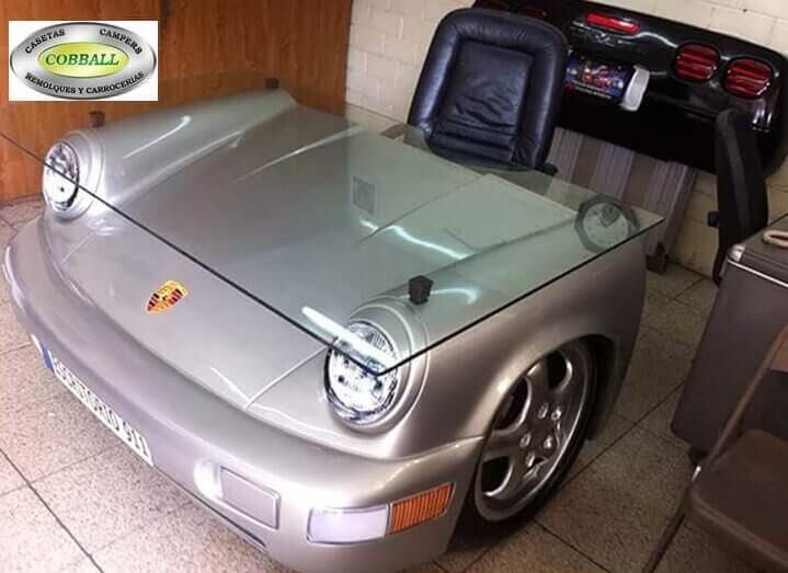 Un escritorio plateado con temática de Porsche, fabricado con la parte delantera de un coche y con una tapa de cristal, situado en un espacio de oficina.