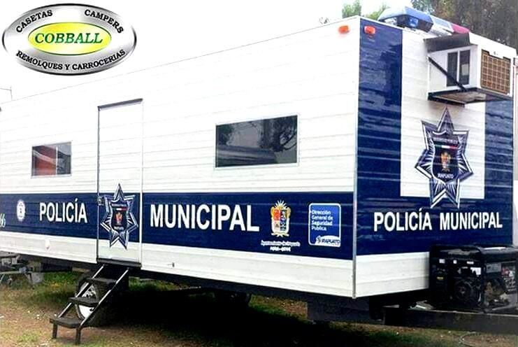 Un remolque de una unidad móvil de la policía municipal, de color blanco y azul, estaba estacionado al aire libre, con emblemas policiales y un aire acondicionado de ventana.