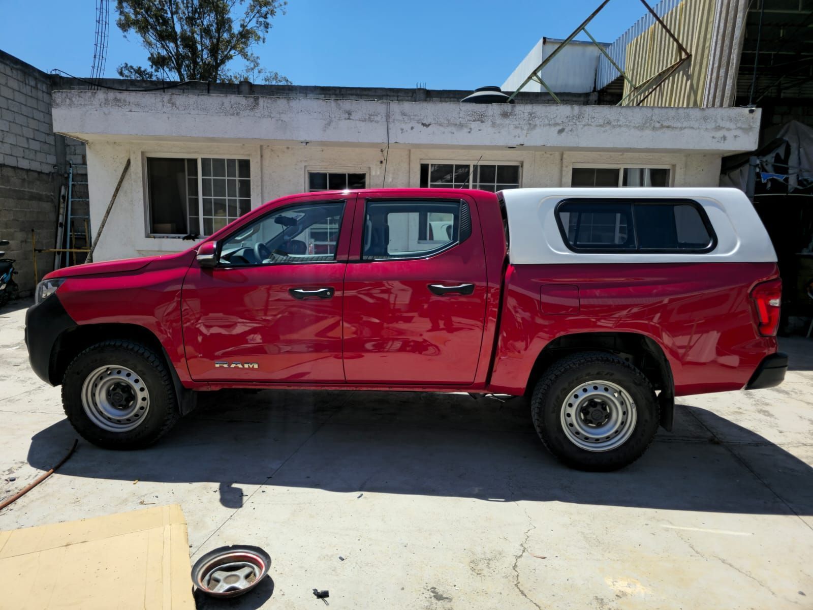 Una camioneta roja de cuatro puertas con una caja de carga blanca estacionada en un terreno de concreto frente a un edificio blanco.