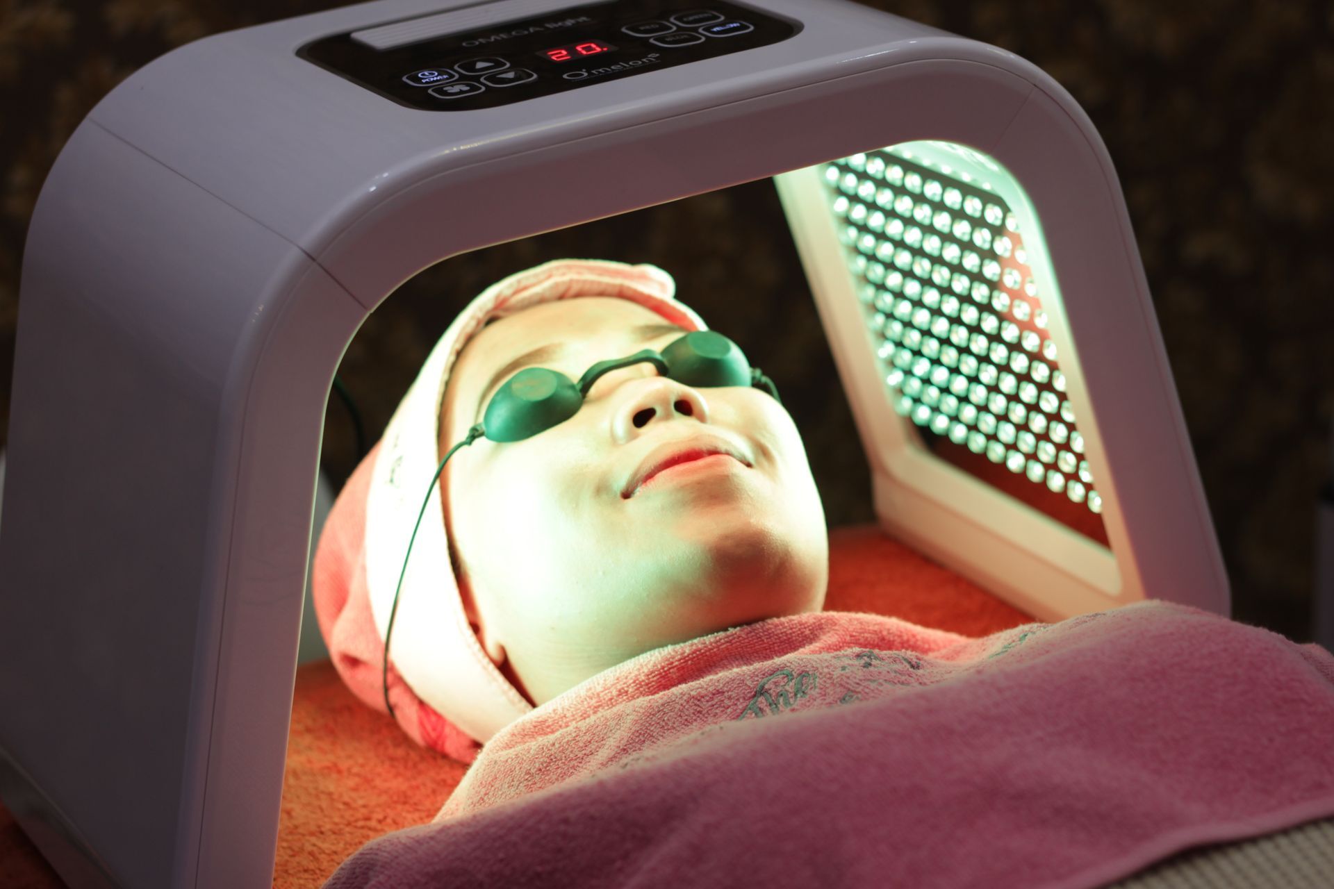 Woman undergoing LED light therapy with protective eyewear in a spa setting, illuminated by red and green lights.