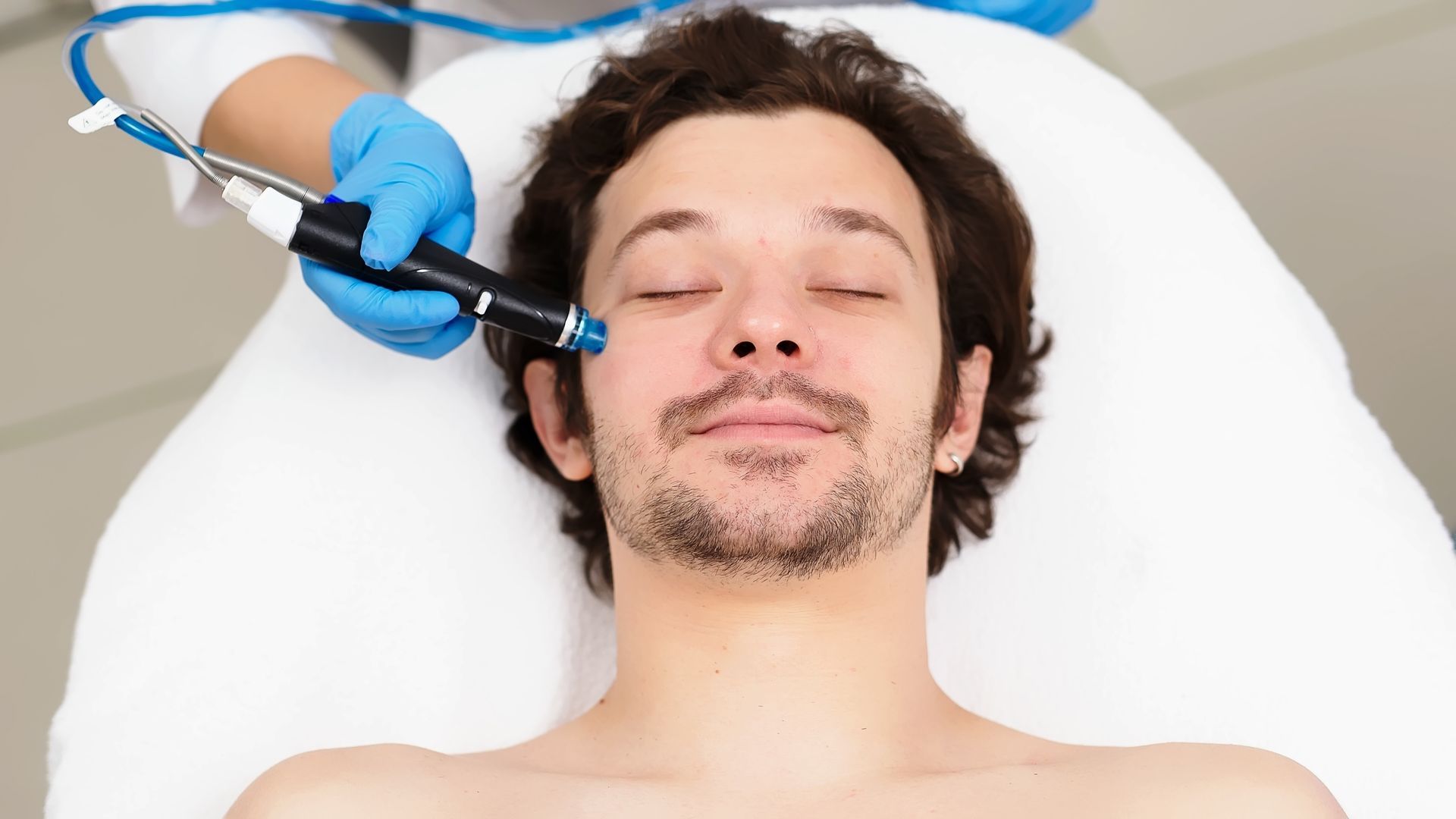 Man receiving facial treatment with a blue-tipped wand held by a gloved hand. He is lying on a white bed with closed eyes.