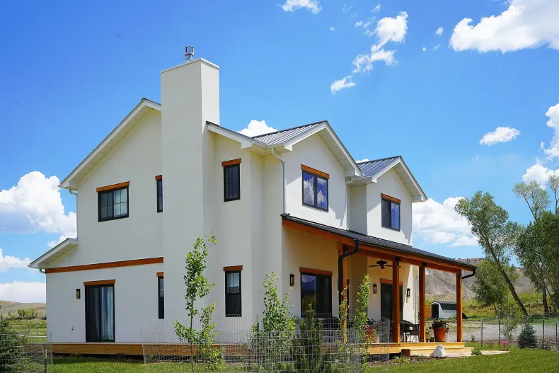 A two-story, cream-colored stucco home with a dark roof and a wooden front porch, set under a blue sky with clouds.