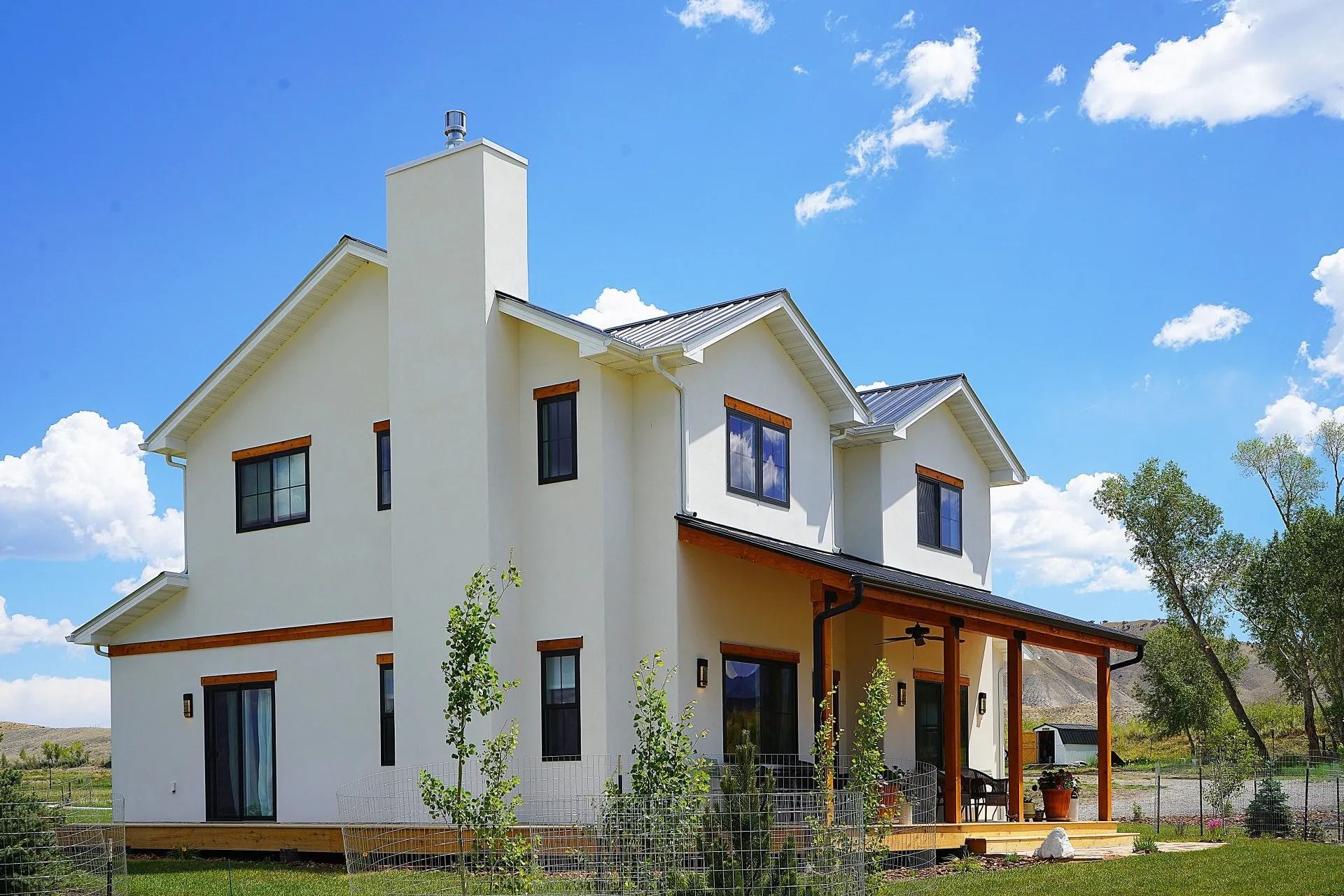 A two-story white house with a tall chimney and a wooden porch under a blue sky with fluffy white clouds.