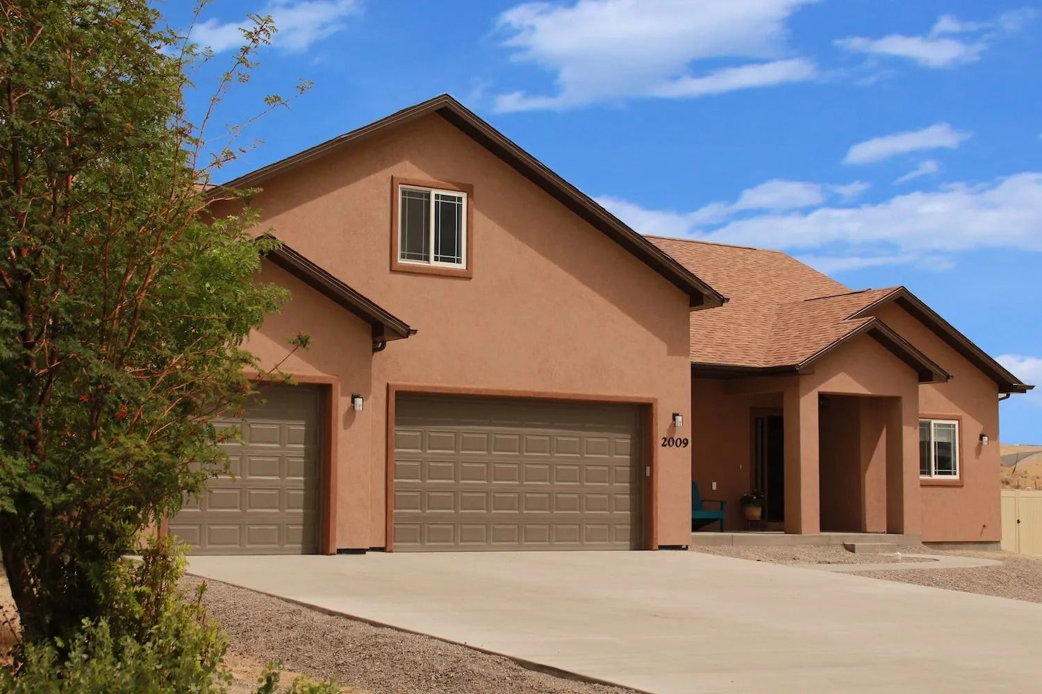A beige, single-story house with a two-car garage, brown roof, and a front entry under a blue sky.