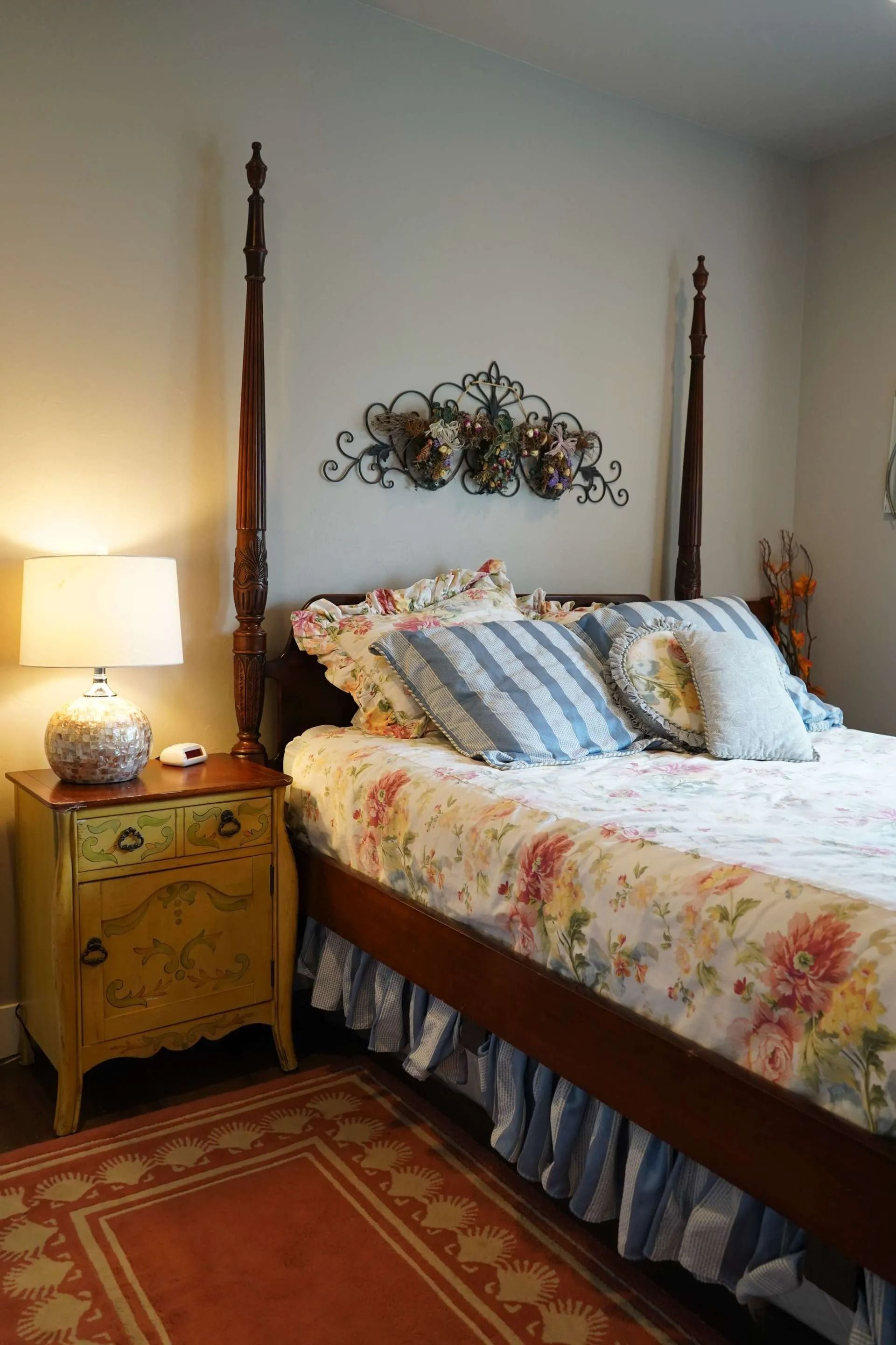A bedroom corner with a wooden four-poster bed featuring floral bedding, a yellow nightstand with a lamp, and a rug.