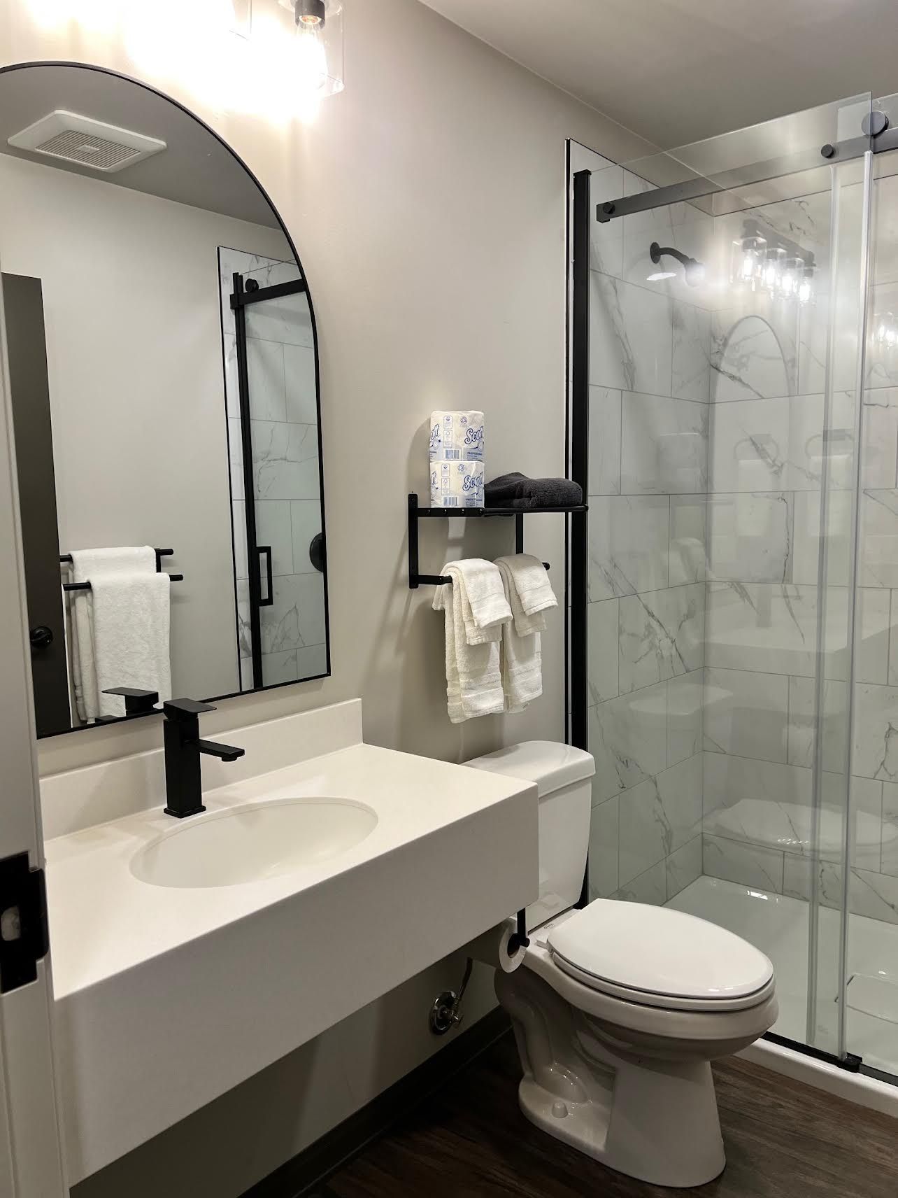 Modern bathroom with white vanity, arched mirror, black accents, and marble-look shower.