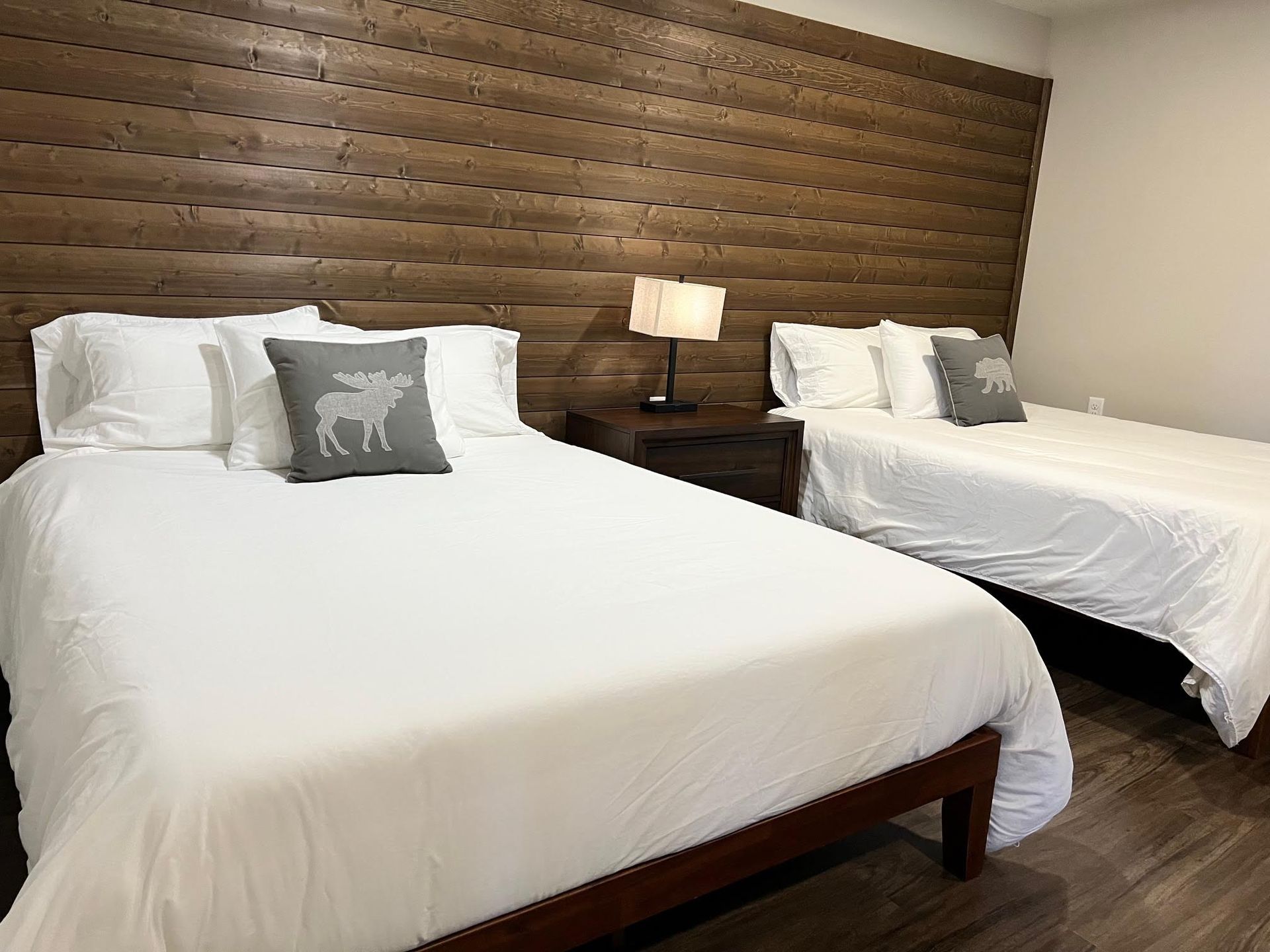 Two beds with white linens, wooden headboard, nightstand with lamp, and grey moose pillows.