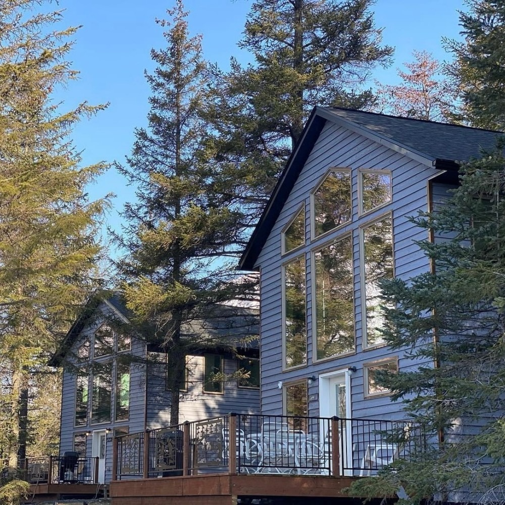 Two-story gray cabin with large windows and a deck nestled among trees under a clear blue sky.