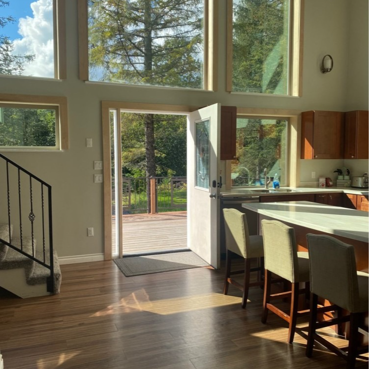 Interior with open doorway to deck, kitchen island with stools, staircase, large windows.