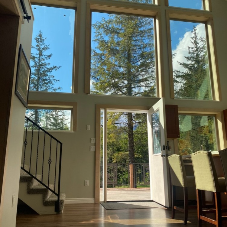 Interior view with large windows overlooking trees and a blue sky; an open door leads to a deck.