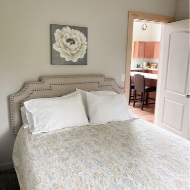 Bedroom with bed, headboard, white pillows, and floral artwork. Open doorway leads to kitchen.