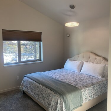 Bedroom with bed, window, and patterned bedding. Neutral walls, gray carpet, and a decorative light.