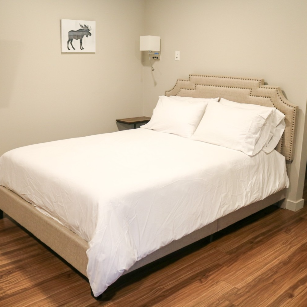 Bedroom with a beige bed and headboard, white bedding, and a small side table. A framed moose picture is on the wall.