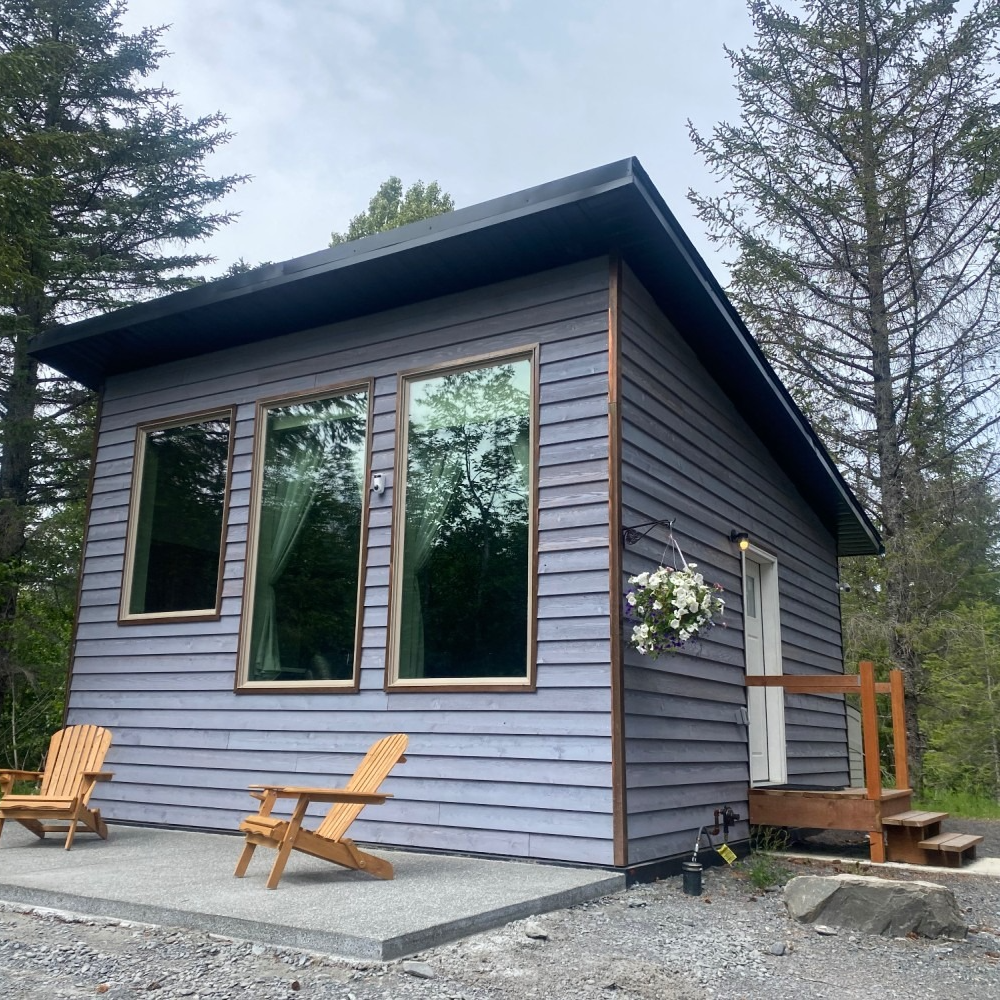 Small gray cabin with large windows and wooden chairs outside.