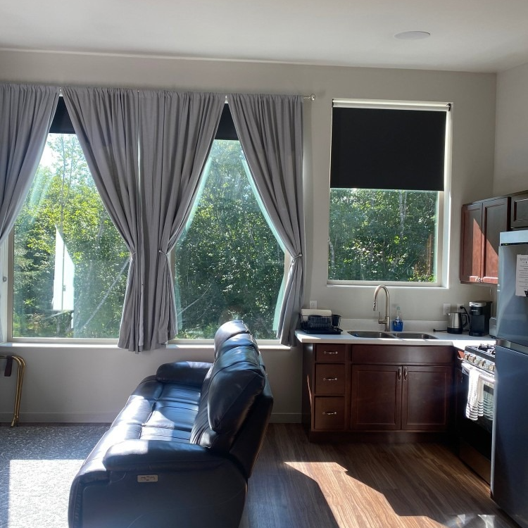 Living room with large windows, gray curtains, dark couch, and a small kitchen with dark cabinets.