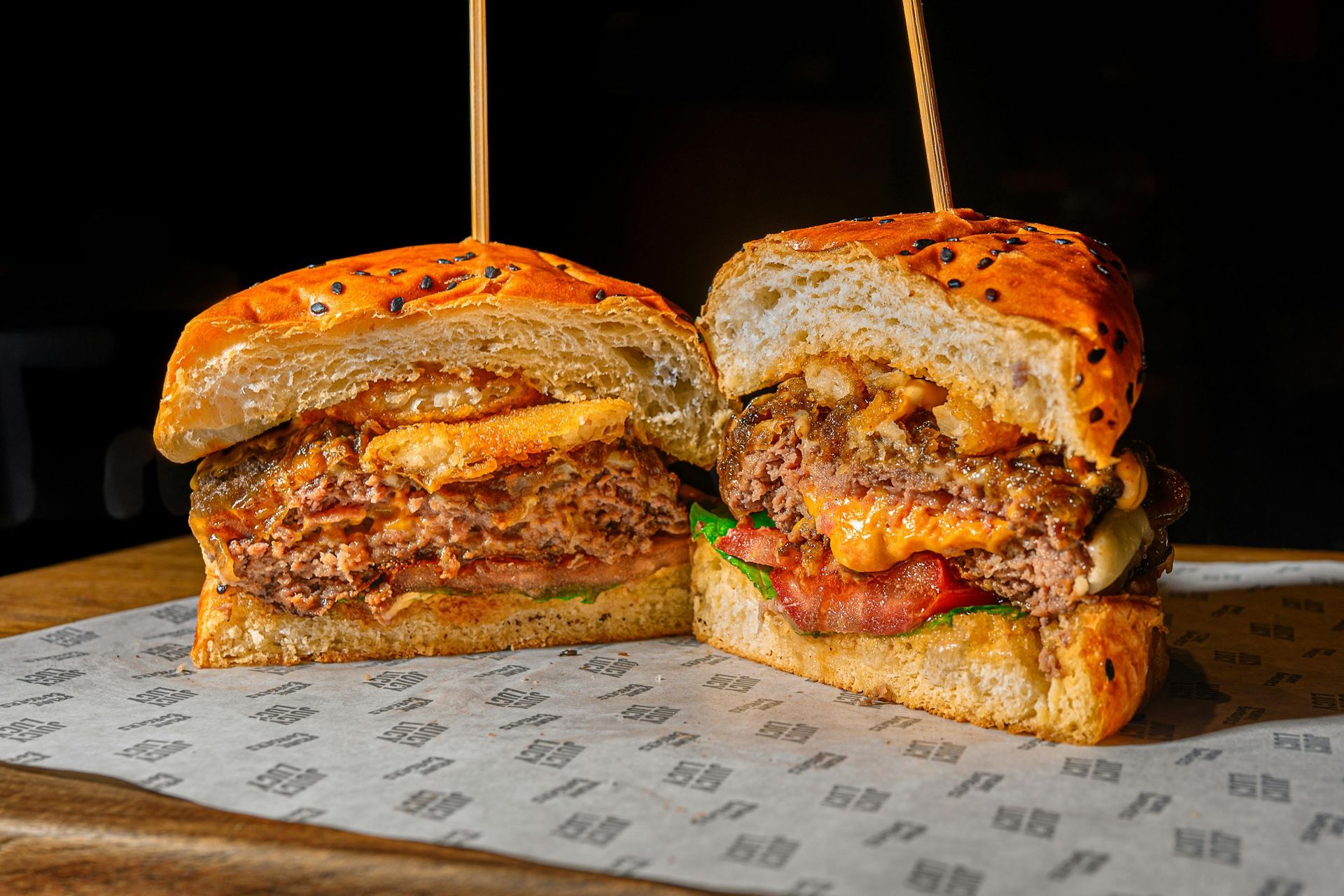 Sliced burger on sesame bun, showcasing a beef patty, bacon, and sauce on patterned paper.