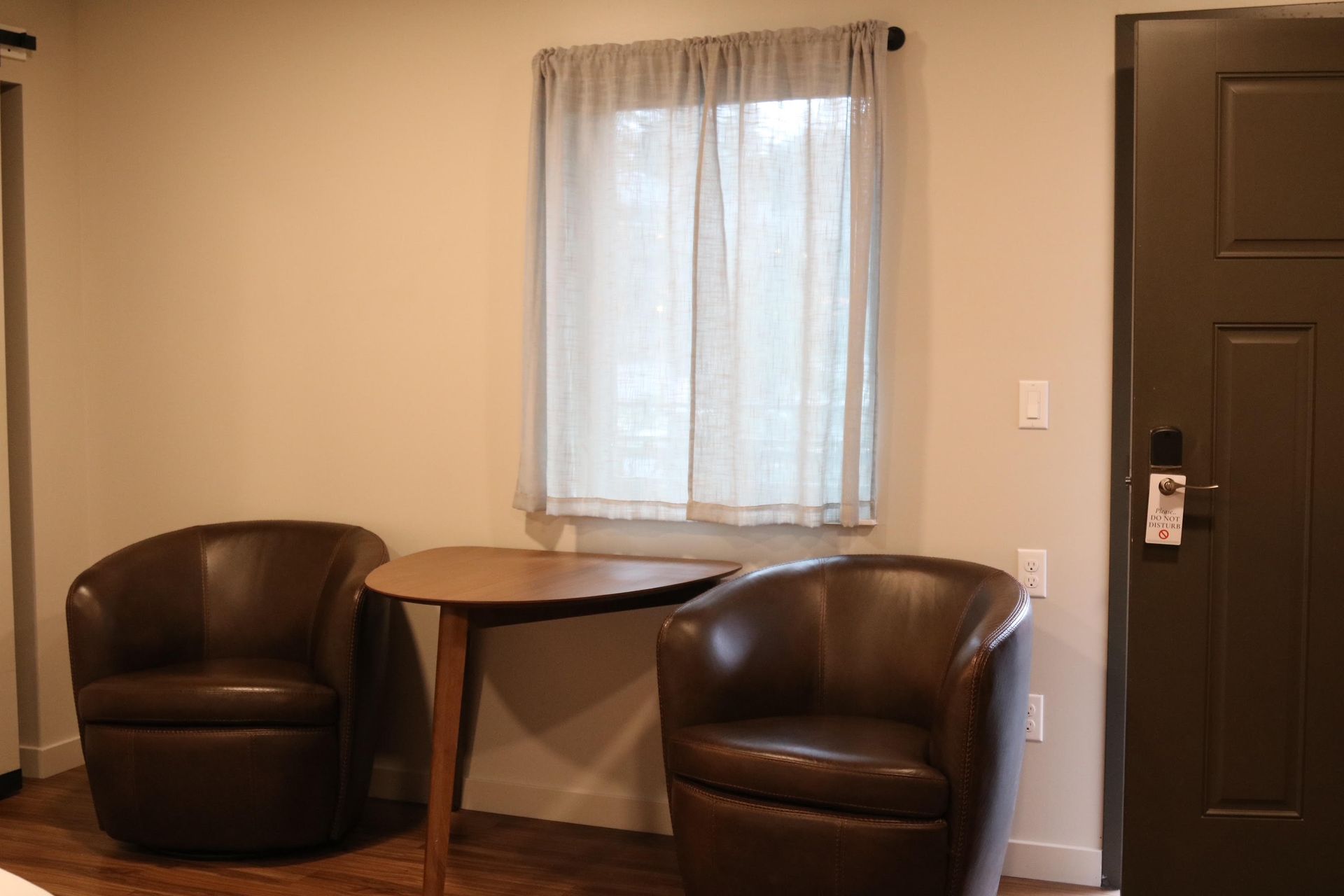 Two brown leather chairs flank a small table under a window with sheer curtains. Dark brown door to the right.