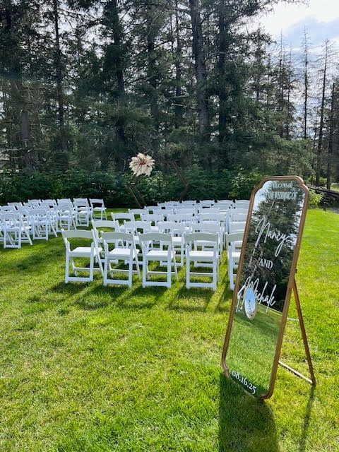 Wedding ceremony setup outdoors with white chairs on grass, a sign, and trees in the background.