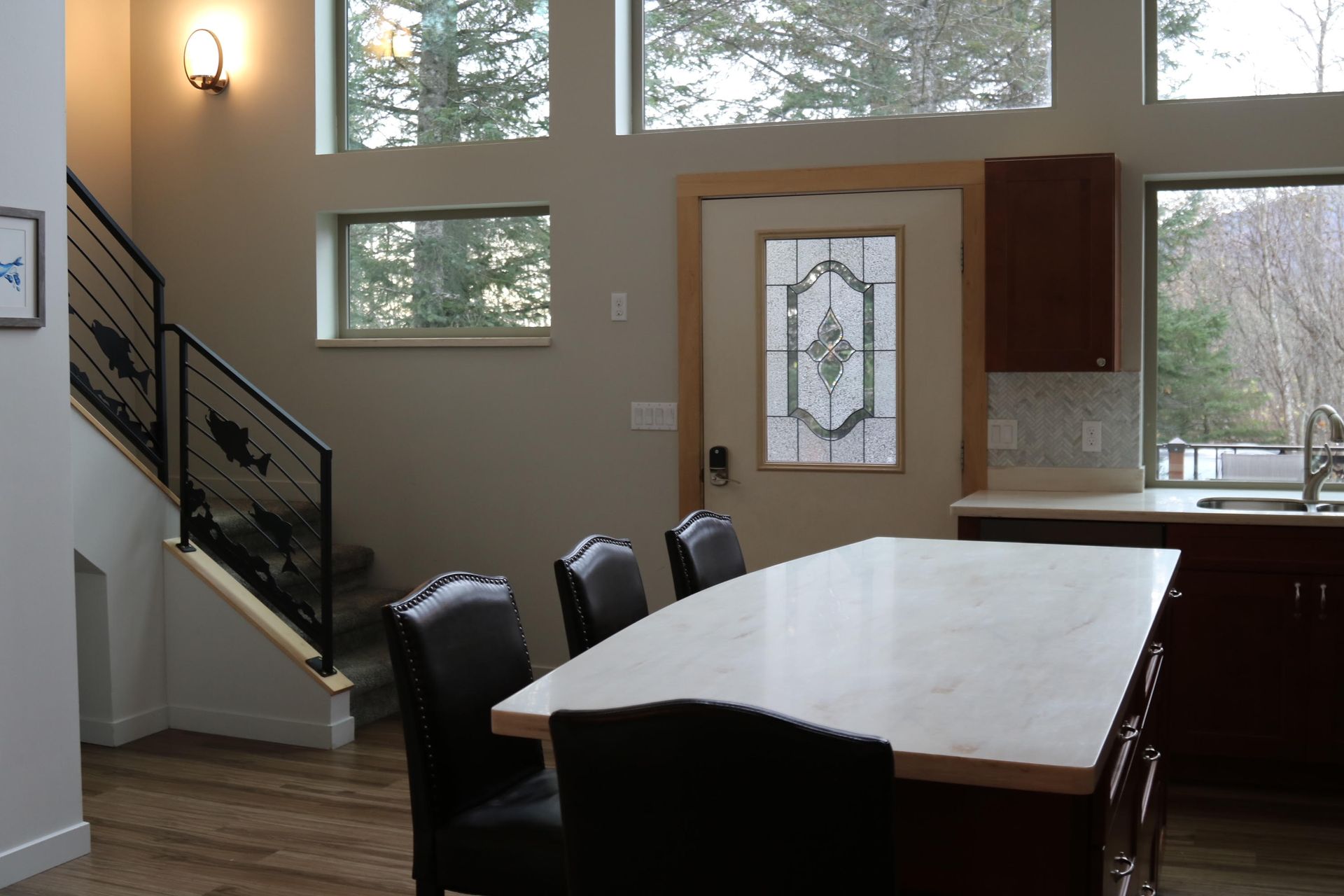 Dining area with table and chairs, stairs, and door. Windows show a view of trees.