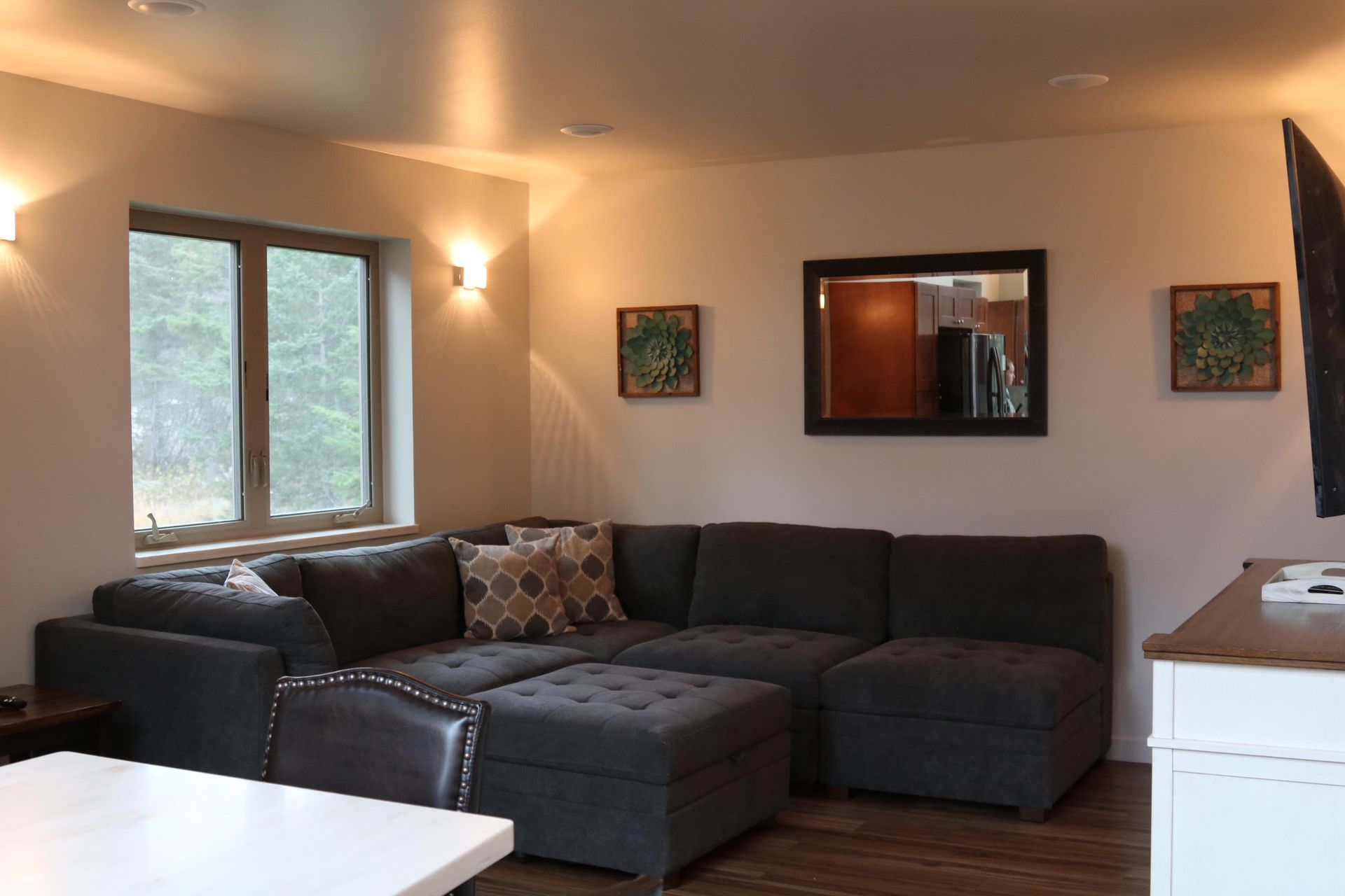 Living room with gray sectional sofa, ottoman, window, art, mirror, and TV.