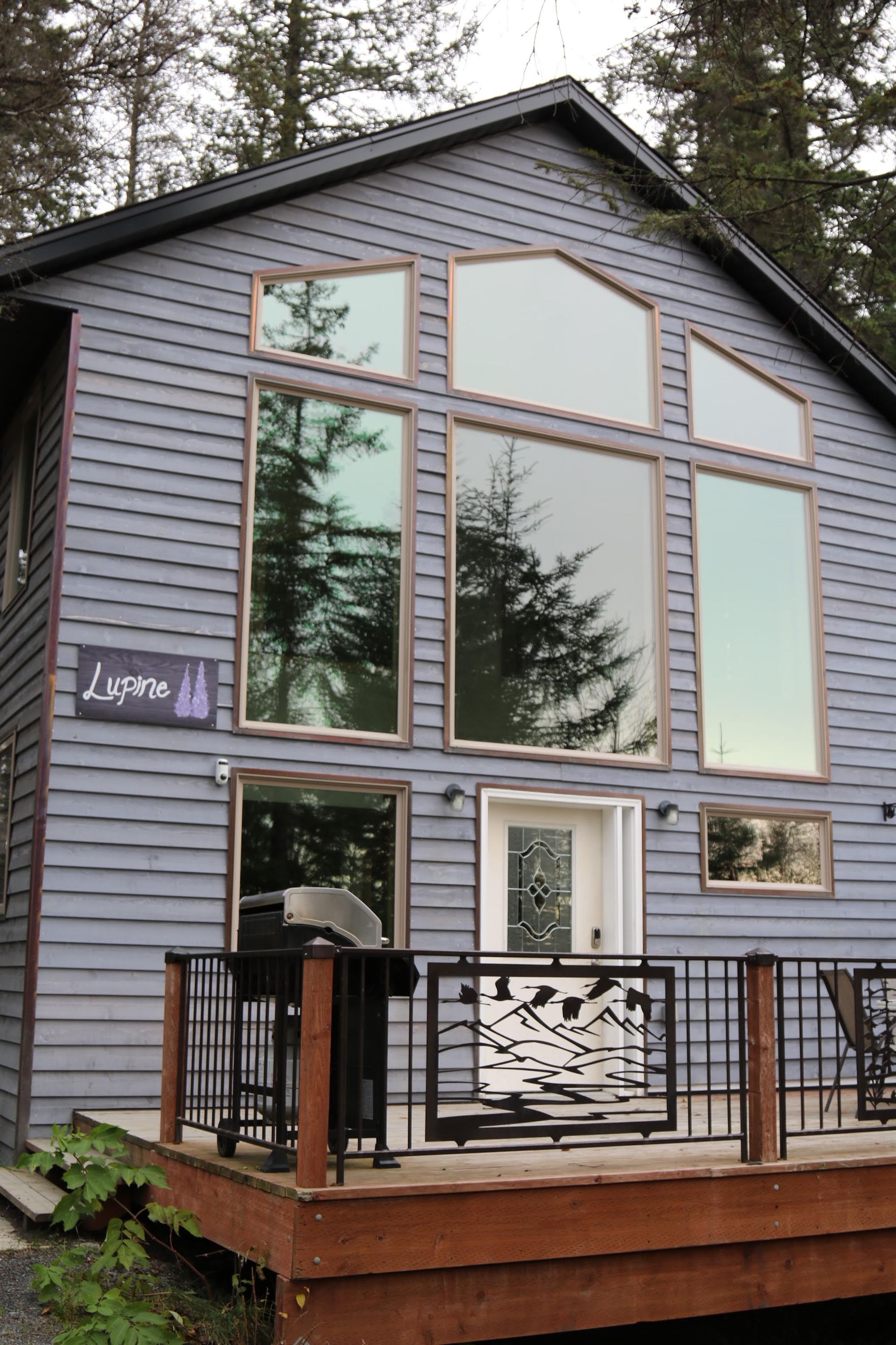 Gray house with large windows, wooden deck, mountain-themed railing.