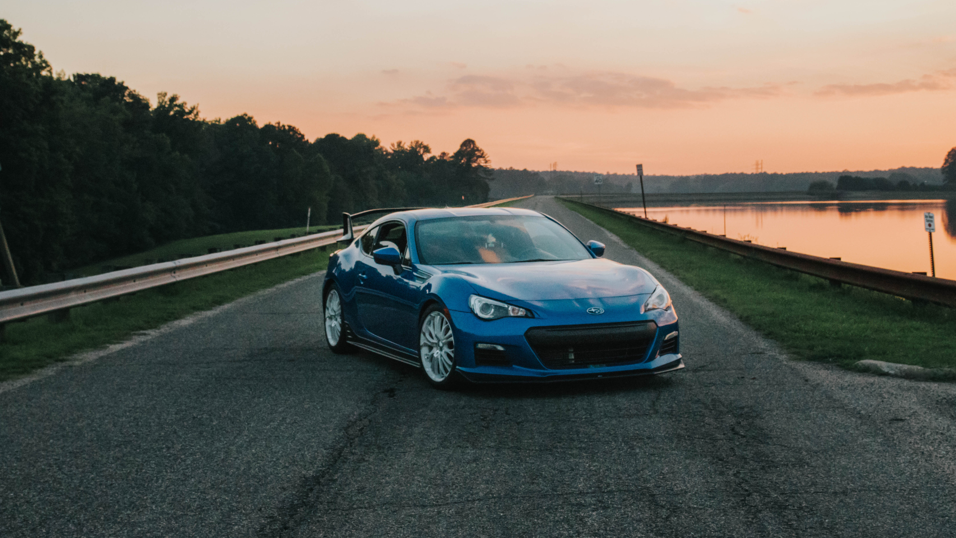 A blue car is driving down a road next to a body of water.