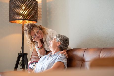 A woman with curly hair smiles while resting her chin on her hand behind another woman seated on a brown leather couch.