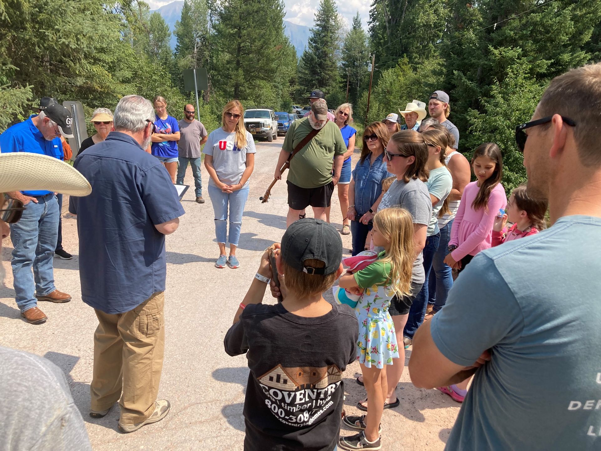 Group of people outdoors, listening to a man speaking. Some are taking photos. Mountains visible in the background.