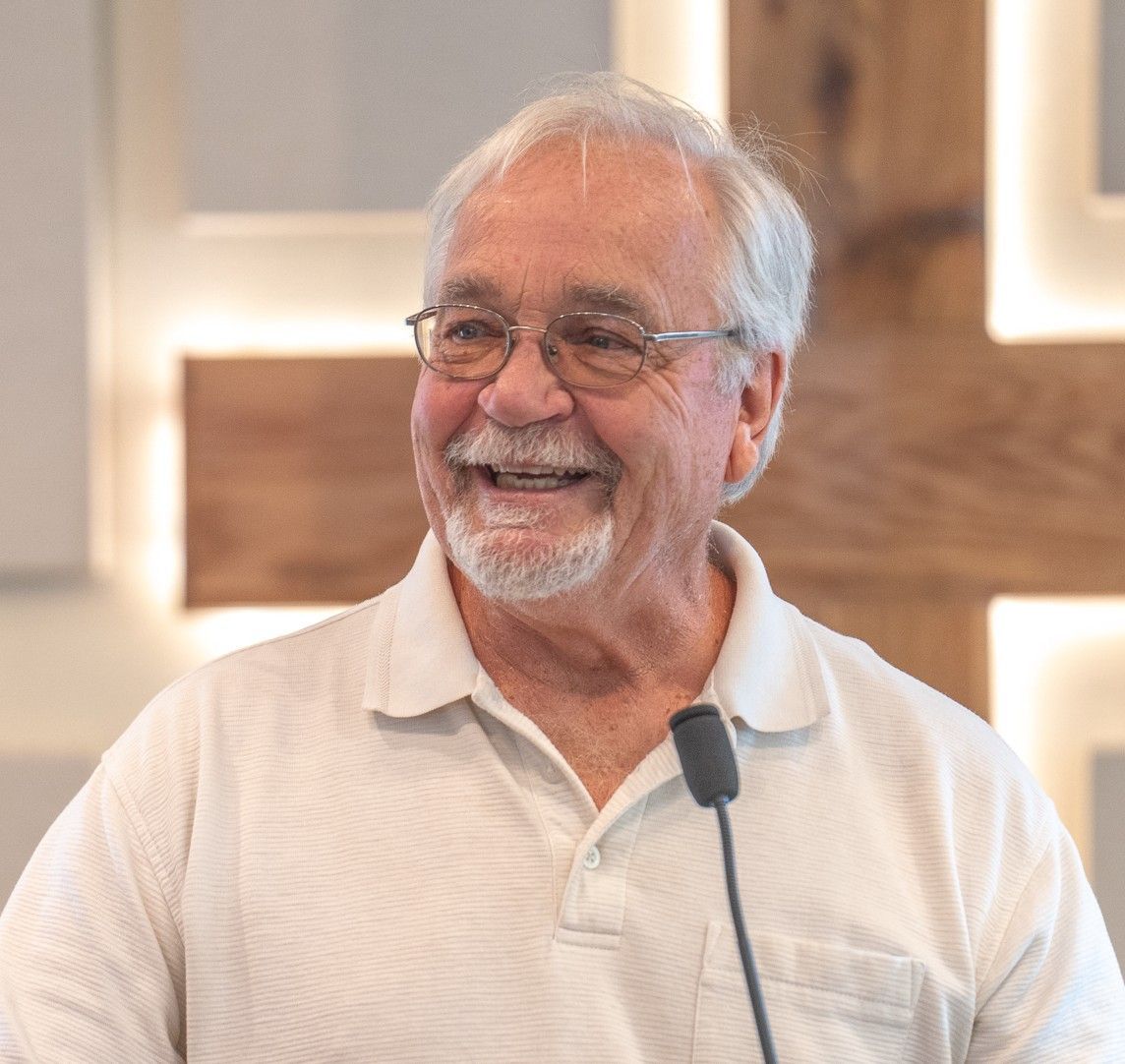 Older man with glasses and beard, wearing bolo tie, in front of a wooden wall.