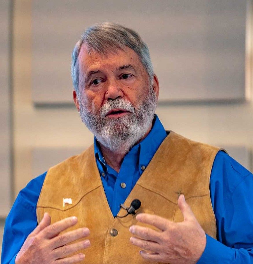 Man in blue shirt and tan vest speaking at a podium, indoors.