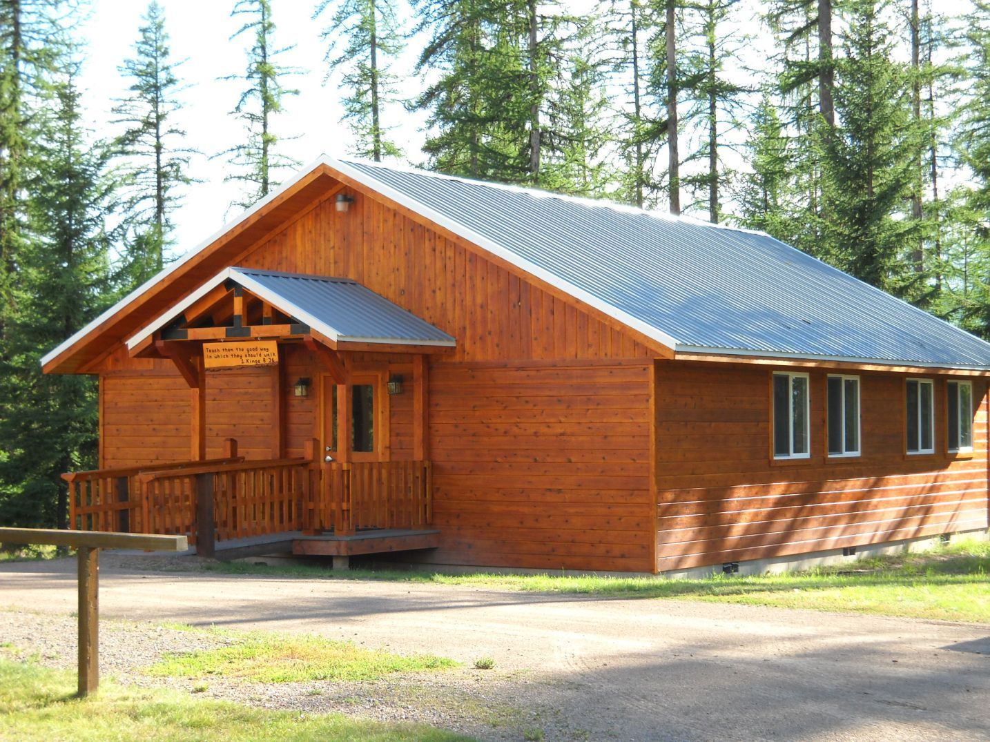 Wooden cabin with gray roof in a forest setting. Front door under small porch, windows on the side.