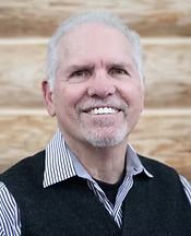 Smiling older man in a black vest and striped shirt against a wood background.