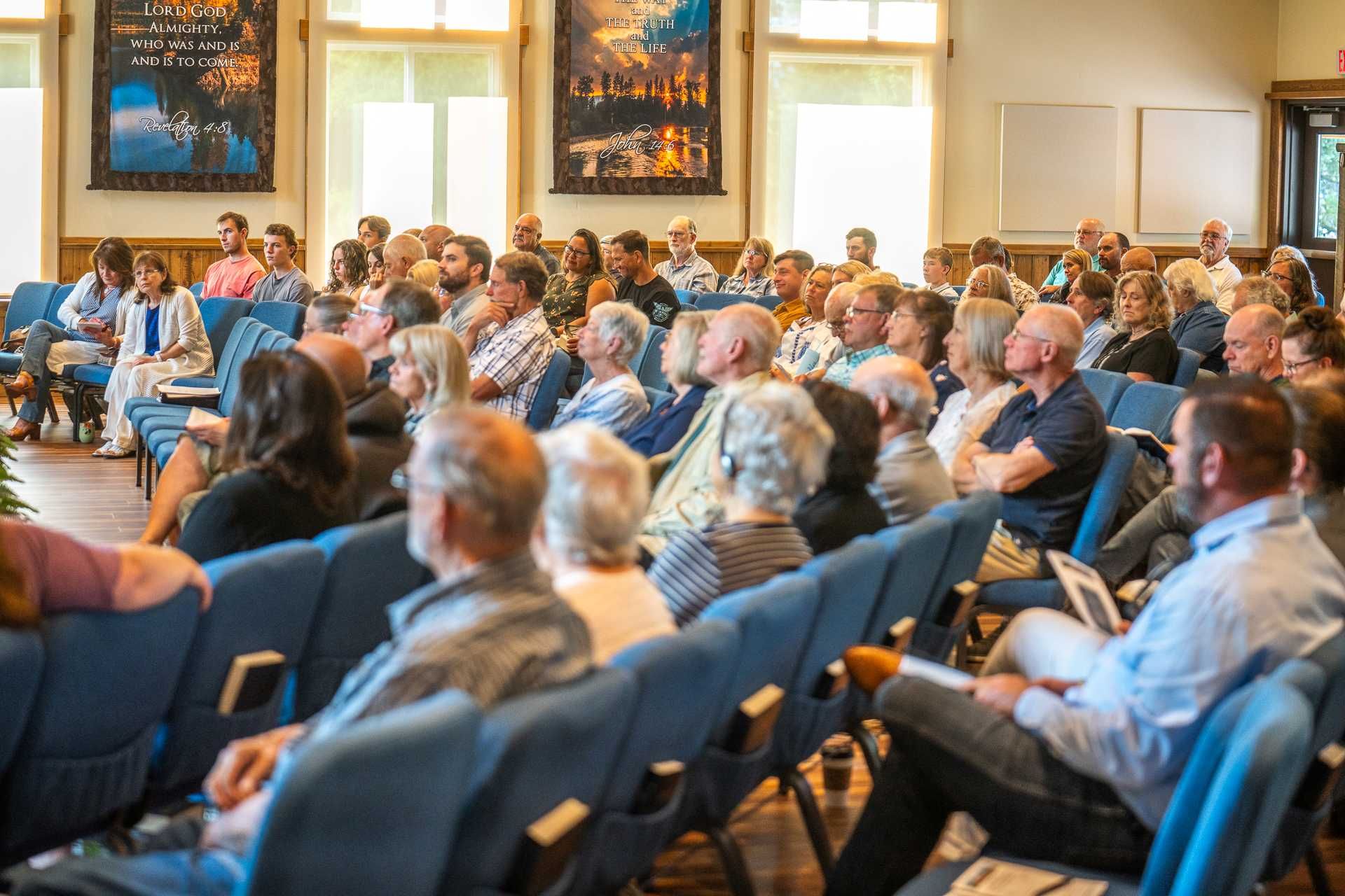 A crowded church service with speakers on a stage and a cross. People are sitting and listening.