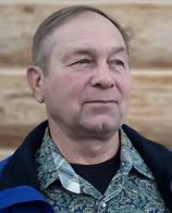Man with light skin, wearing a patterned shirt, black top, and blue jacket. He has a neutral expression, outdoors.