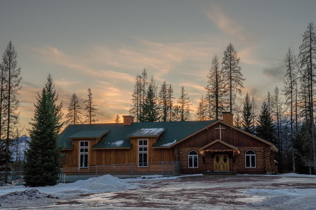 Log cabin in snow, surrounded by trees, under a sunset sky.