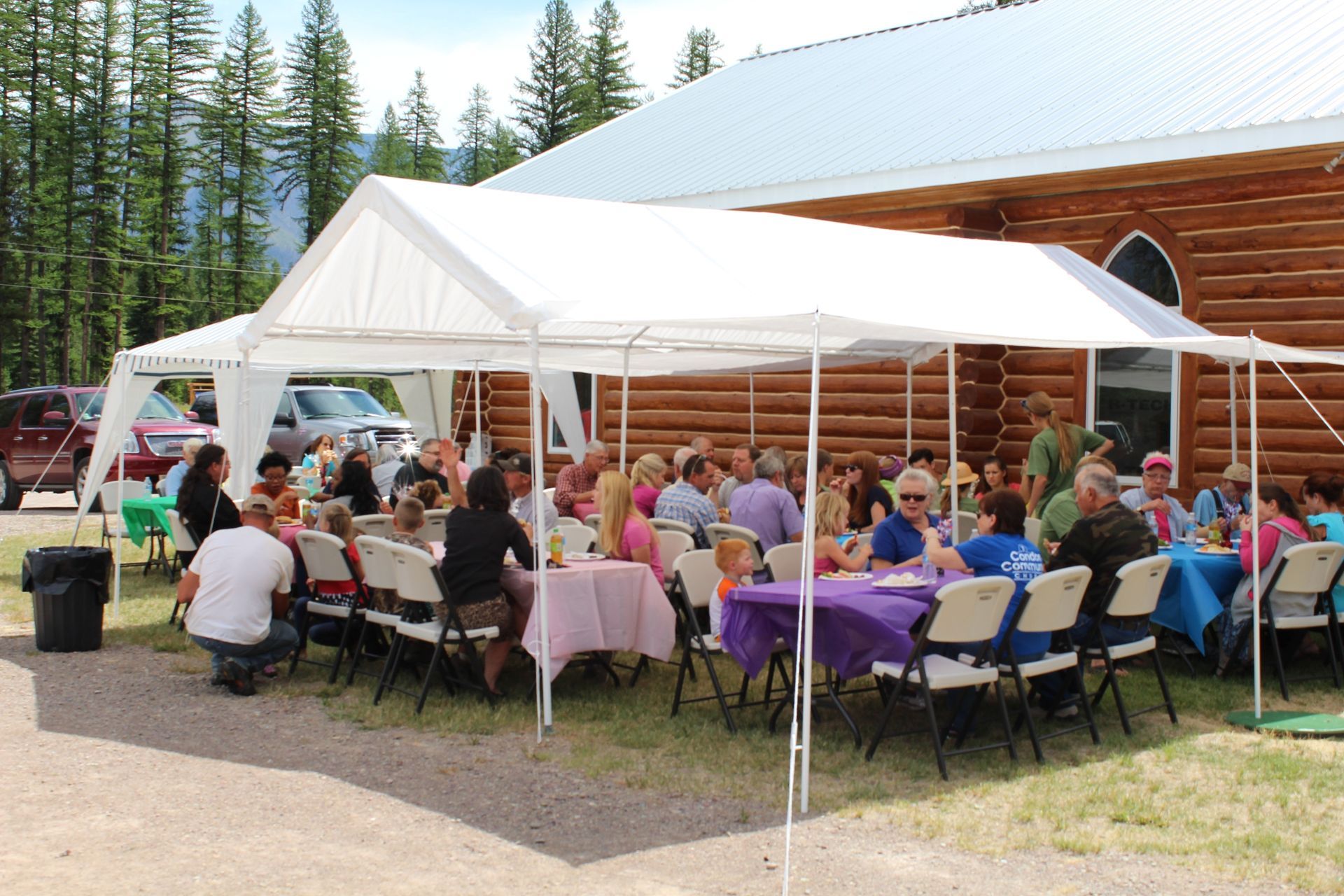 People gathered under a white tent, seated at tables, by a log cabin church.