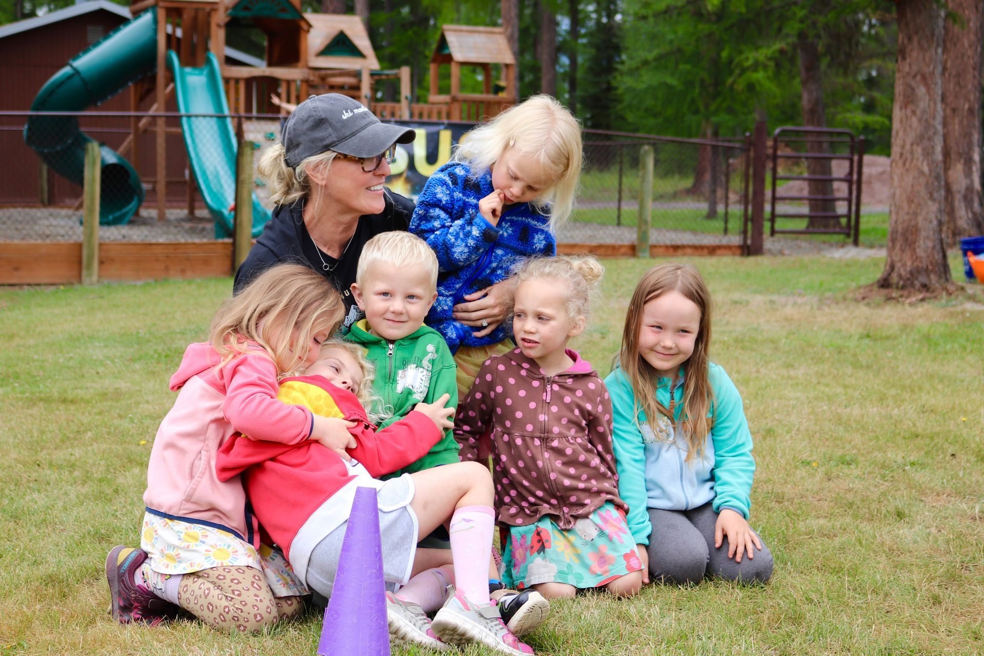 A group of children and a woman smile together on a grassy playground.