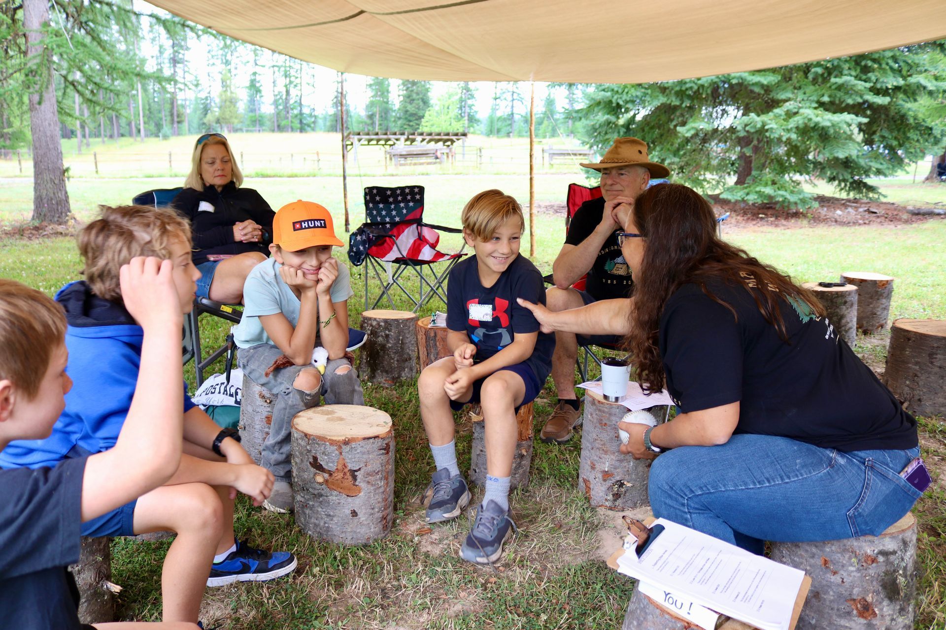 Group of people, including children, seated outdoors under a canopy, engaged in conversation.