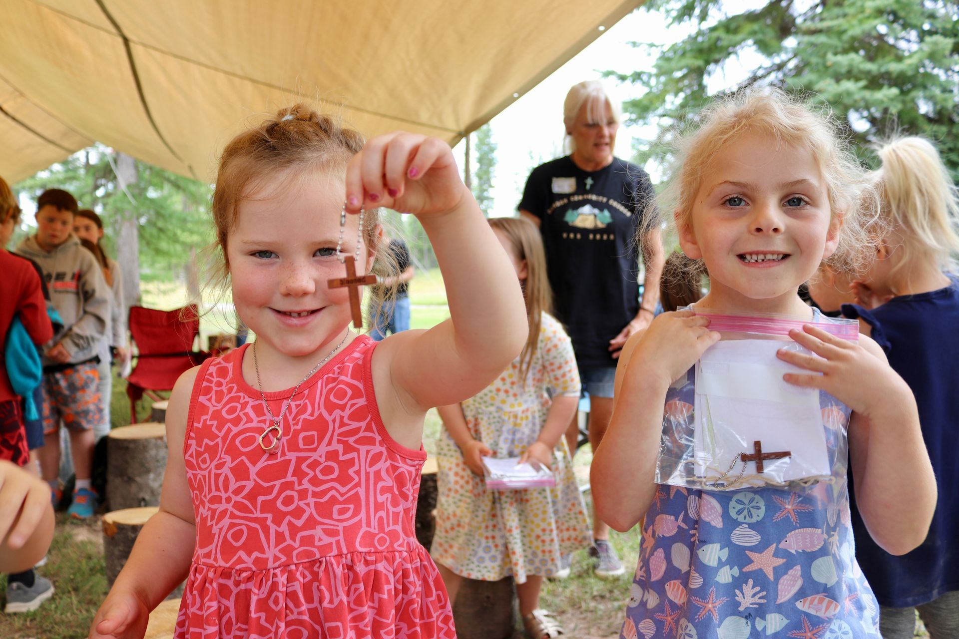 Two young girls holding up wooden crosses, smiling, at an outdoor event, with a woman in the background.