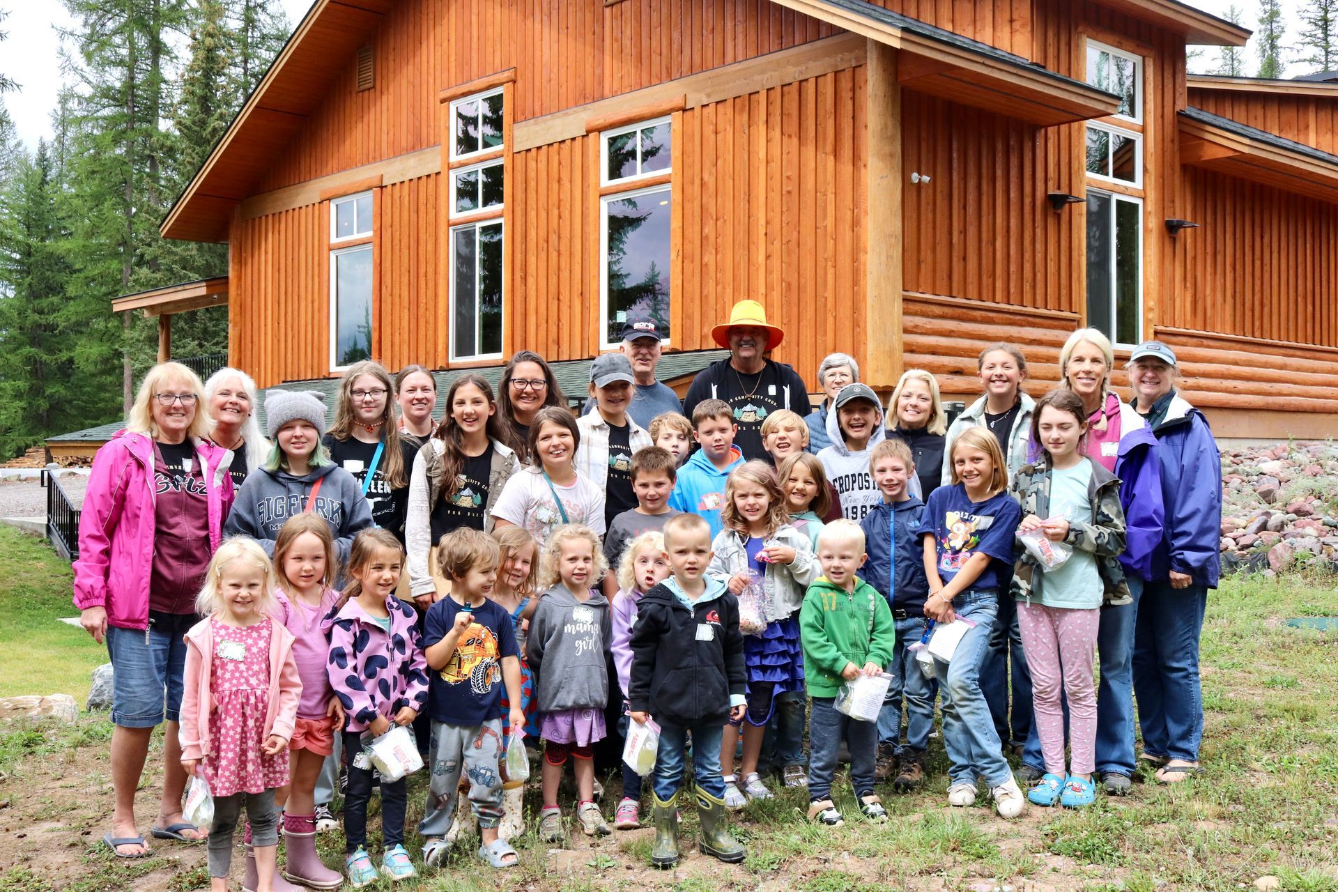 Group of children and adults posing in front of a wooden lodge. Some children hold small trees.