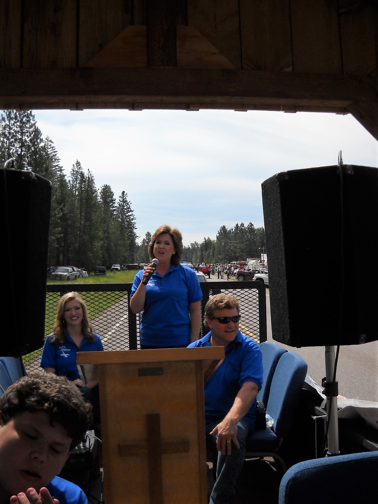 Woman speaking at a podium outdoors, with two other people in blue shirts nearby. A cross is on the podium.