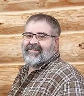 Man with glasses and a grey beard smiles in front of a wooden wall, wearing a plaid shirt.