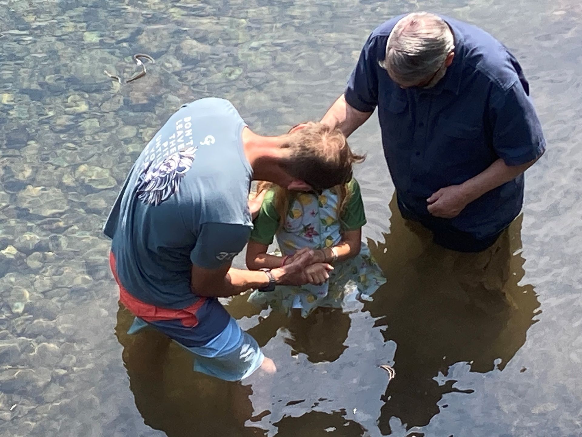 Man baptizing a girl in a shallow river, another man assisting.